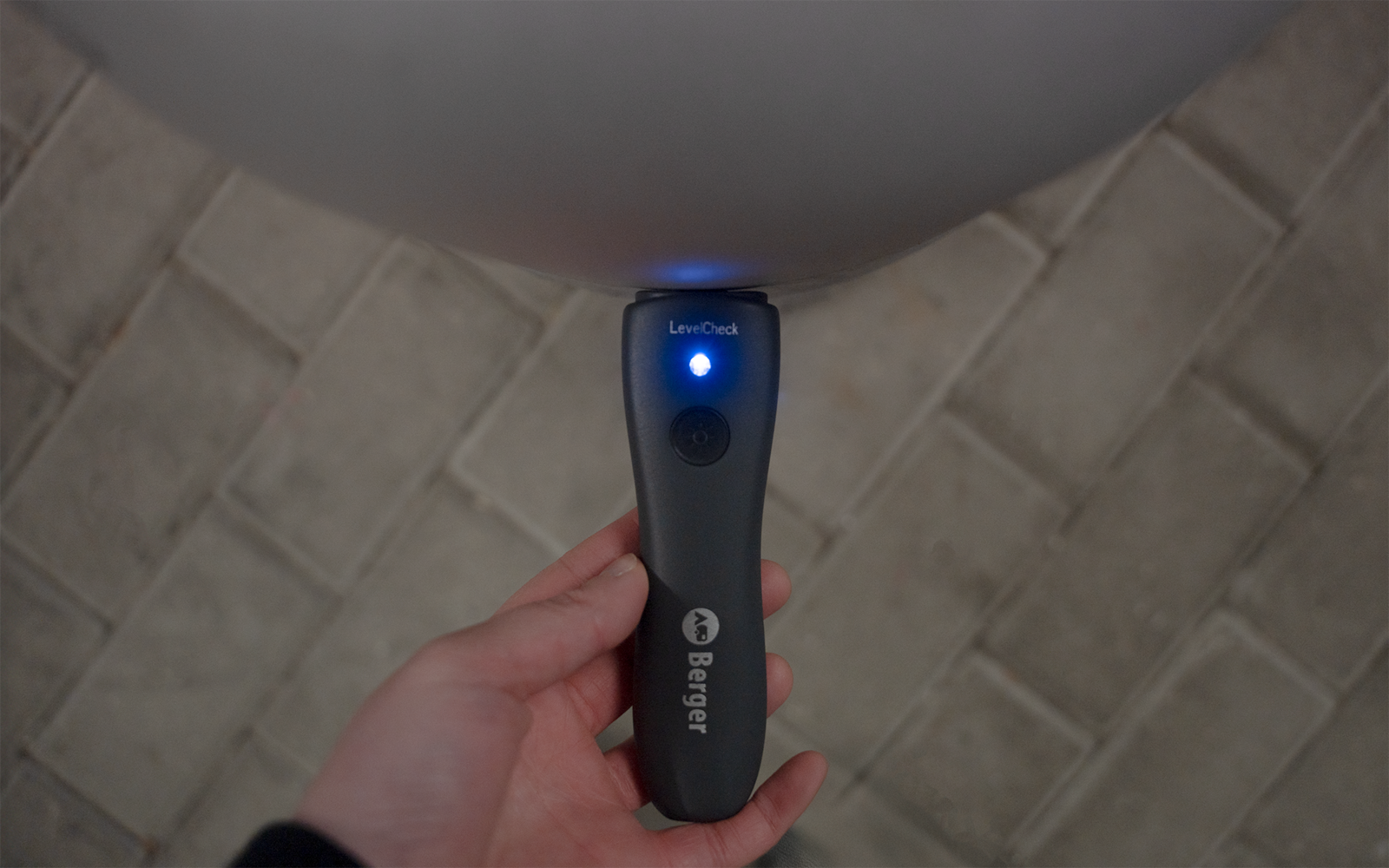 Hand hält ein schwarzes Handgerät mit blau leuchtender Diode, einem Knopf und weißer Beschriftung; darüber ein rundes graues Objekt; Boden aus rechteckigen Pflastersteinen