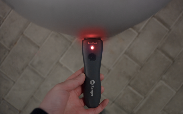 Hand hält ein schwarzes Handgerät mit roter LED oben, rundem Knopf und sichtbarem Schriftzug 'Energizer'; im Hintergrund gefliester Boden und ein rundes weißes Objekt