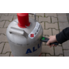 Silver gas cylinder with red valve guard, hazard label 'UN 1965' and printed 'PROPAN', standing on paving; a person's hand holds a small device with a green light against the cylinder
