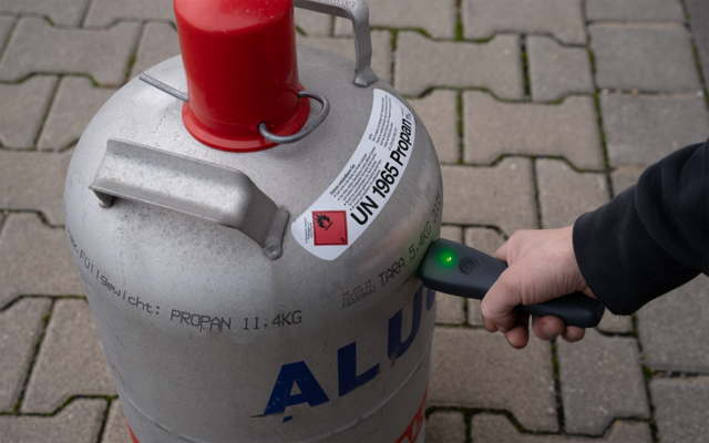 Silver gas cylinder with red valve guard, hazard label 'UN 1965' and printed 'PROPAN', standing on paving; a person's hand holds a small device with a green light against the cylinder