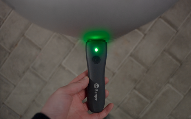 Hand holding a black elongated device with a green light dot at the top and a button below; background: light floor with rectangular tiles