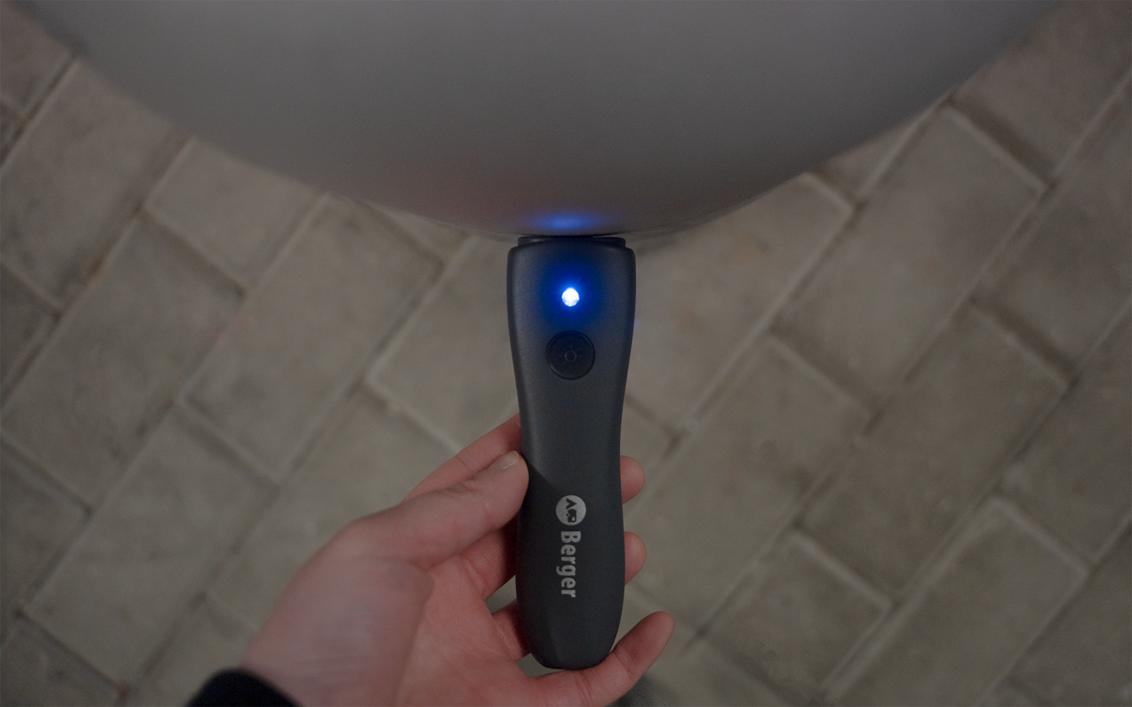 Hand holding a black handheld device with a blue lit LED, a button and white markings; above it a round gray object; floor of rectangular paving stones