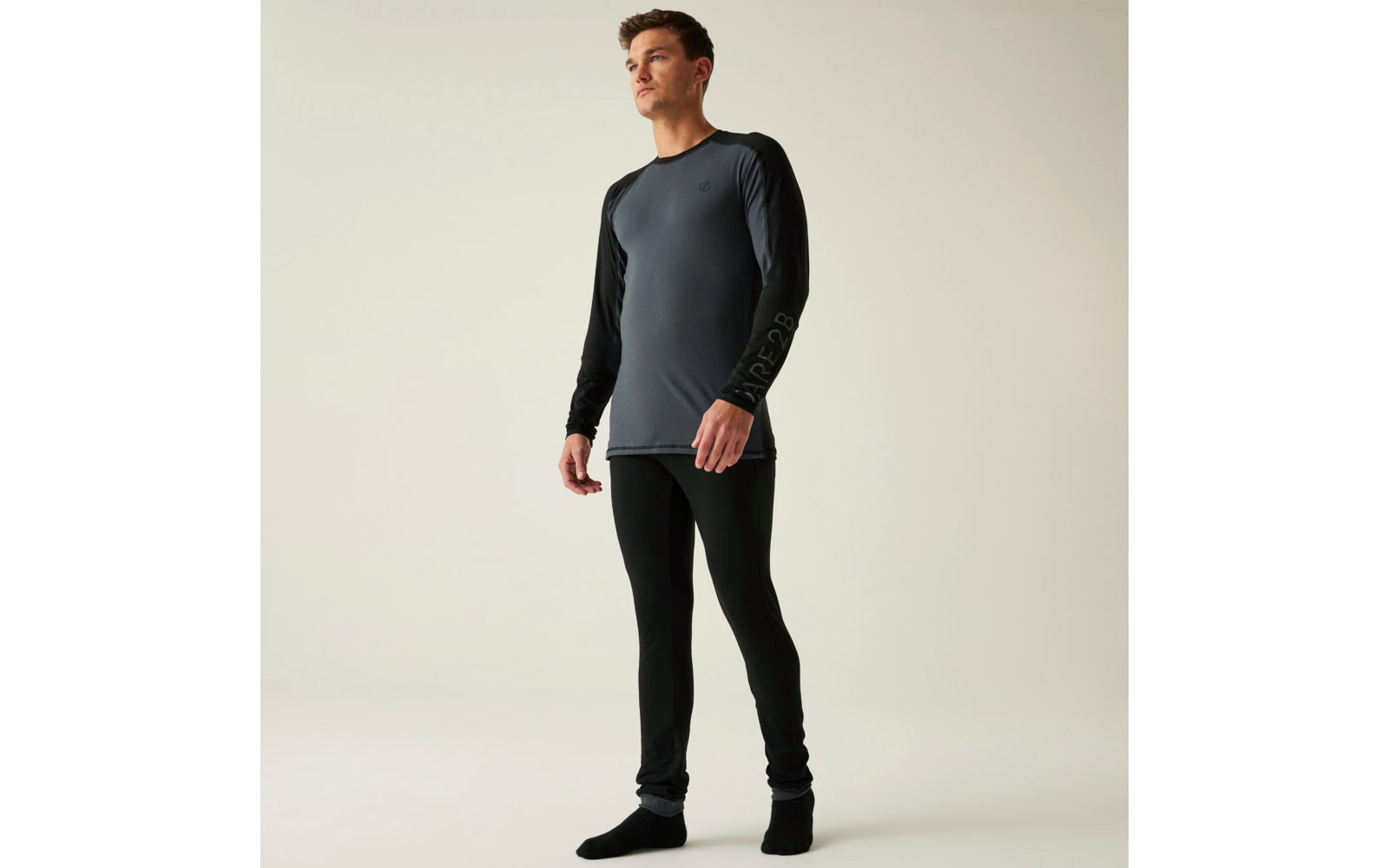 Stehende Person in grauem Langarmshirt mit schwarzen Ärmeln, schwarzen Leggings und schwarzen Socken vor hellem einfarbigem Hintergrund