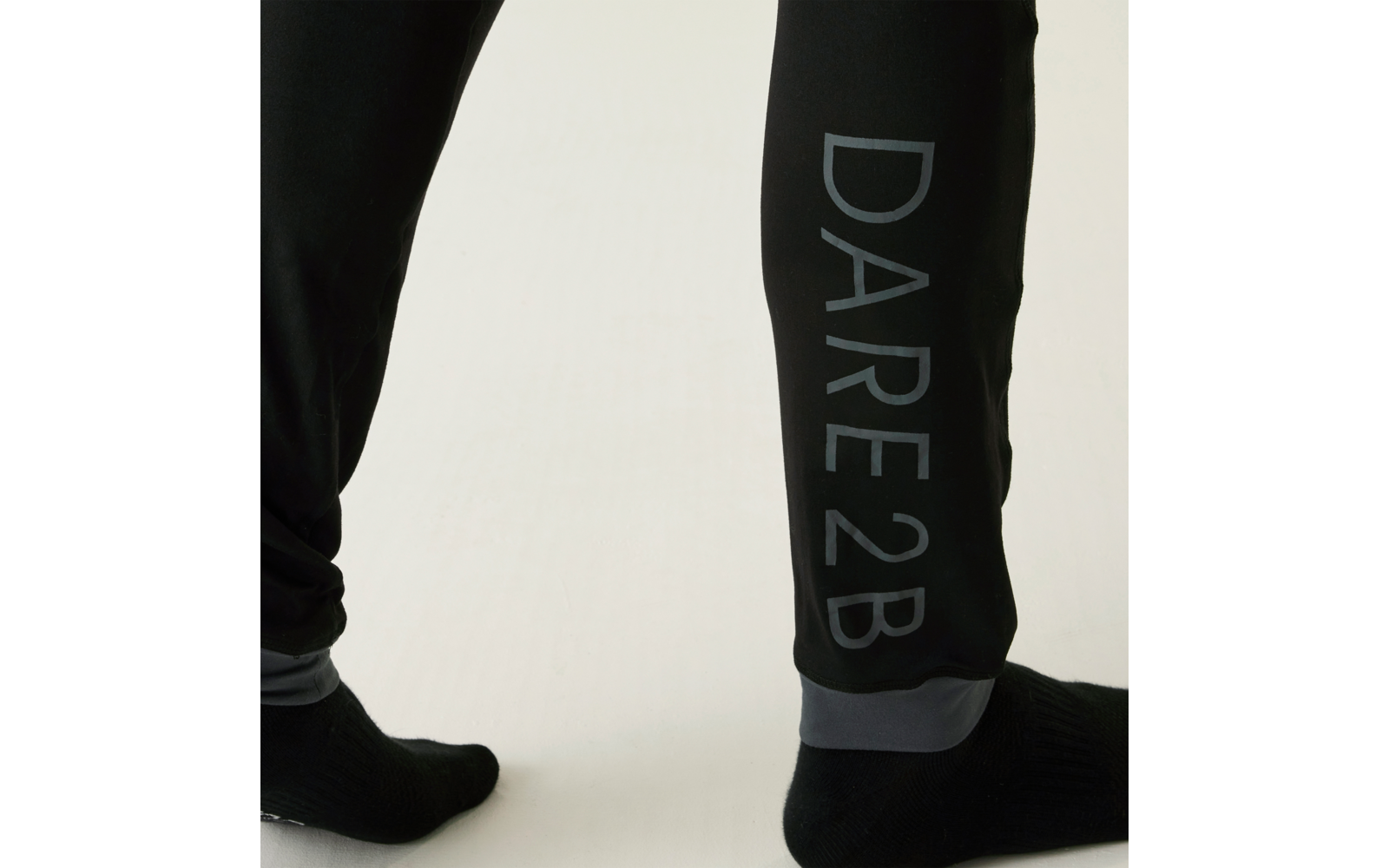 Untere Beine in schwarzen Jogginghosen, Hosenbein mit senkrechter Aufschrift "DARE2B", schwarze Socken, heller Hintergrund