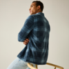 Man zit zijwaarts op een houten stoel, rug zichtbaar, draagt een blauw geruit overhemd en lichte spijkerbroek, kijkt naar links