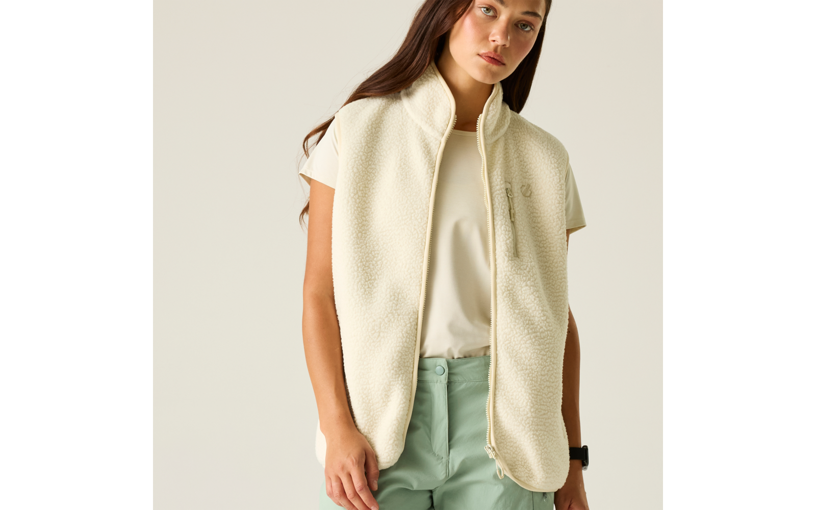 Personne visible de la mi-cuisse vers le haut, porte une veste sans manches en polaire crème avec fermeture éclair à moitié ouverte, T‑shirt blanc à manches courtes, pantalon vert clair, cheveux longs bruns, montre noire au poignet gauche, fond uni clair