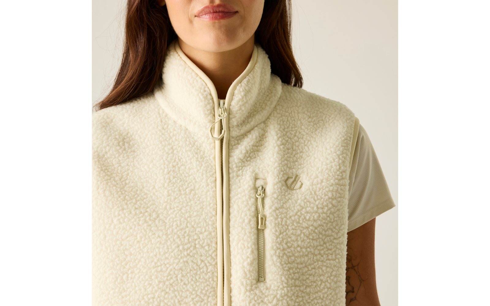 Torse et cou d'une personne portant une veste de couleur crème à col montant et fermeture éclair devant, poche poitrine zippée visible, t-shirt à manches courtes en dessous, partie inférieure du visage visible (menton et lèvres), cheveux bruns, tatouage sur le haut du bras