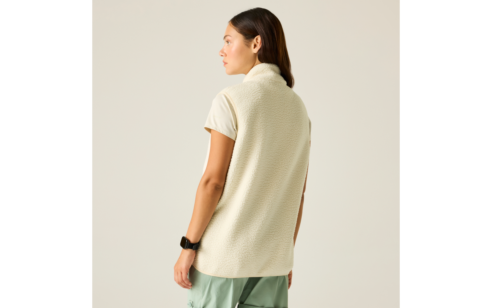 Personne vue de dos, porte une veste sans manches en polaire crème sur un T‑shirt beige à manches courtes, pantalon vert clair et montre connectée au poignet gauche, debout sur fond beige, regarde vers la gauche
