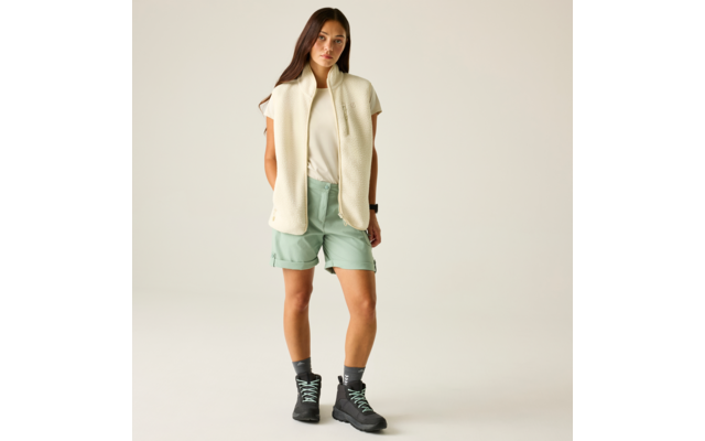 Femme debout sur fond neutre, porte un gilet sans manches crème, un T‑shirt blanc, un short vert menthe et des chaussures de randonnée grises avec lacets vert clair