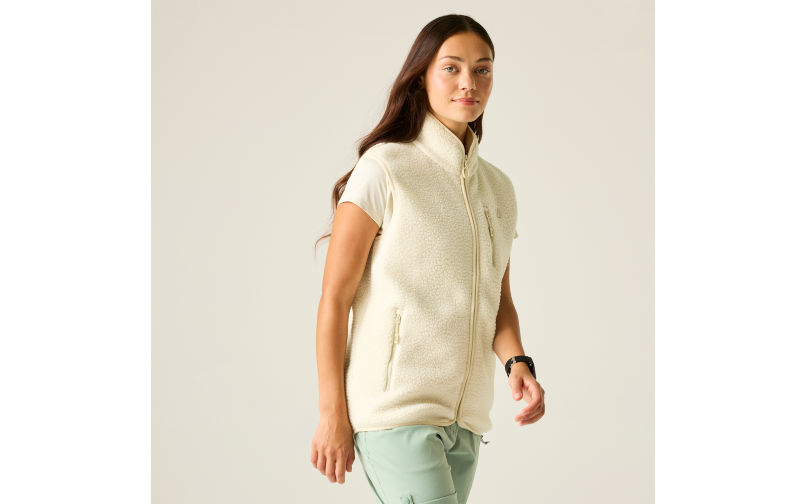 Femme en gilet polaire crème, T‑shirt à manches courtes et pantalon vert clair, marchant vers la droite sur un fond neutre et clair, portant une montre