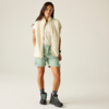 Femme debout sur fond neutre, porte un gilet sans manches crème, un T‑shirt blanc, un short vert menthe et des chaussures de randonnée grises avec lacets vert clair