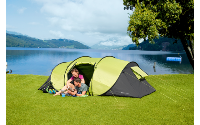 Una mujer y dos niños sentados frente a una tienda verde sobre la hierba junto a un lago