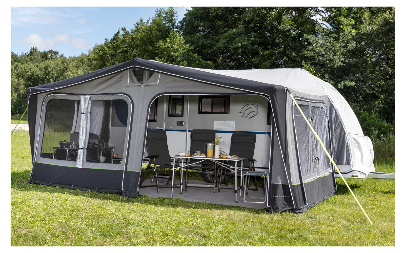 Large gray awning with open windows and a table with chairs in front, on green grass, trees in the background