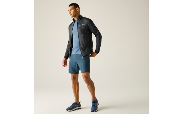 Person steht, trägt dunkle Jacke, hellblaues T‑Shirt, türkis-blaue Shorts und blaue Sportschuhe; Blick nach links; heller einfarbiger Hintergrund