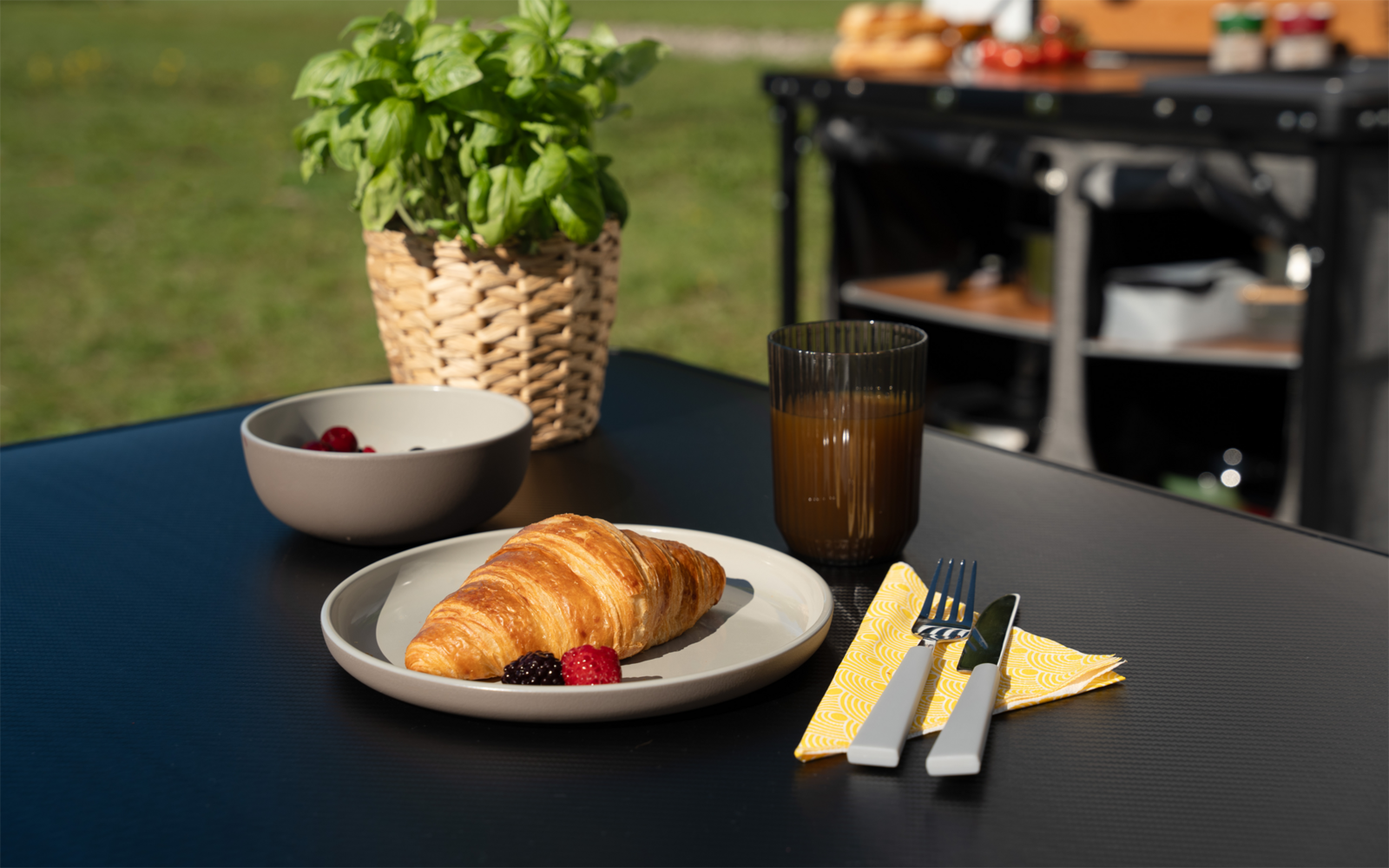 Croissant auf weißem Teller mit zwei Himbeeren, kleine weiße Schale mit Himbeeren, braunes Glas mit Saft, Messer und Gabel auf gelber Serviette, Basilikum in geflochtenem Korb auf schwarzem Tisch, im Hintergrund Rasen und ein Gartenwagen