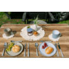 Houten tafel met twee gedekte plaatsen: links een licht bord met een broodje en groene druiven, rechts een blauw bord met een broodje en rode druiven; vorken links, messen en lepels rechts; twee mokken op gehaakte onderzetters; in het midden een gehaakt kleedje met een jampot, een witte keramische ananas en een oranje vrucht; gras en autowiel op de achtergrond