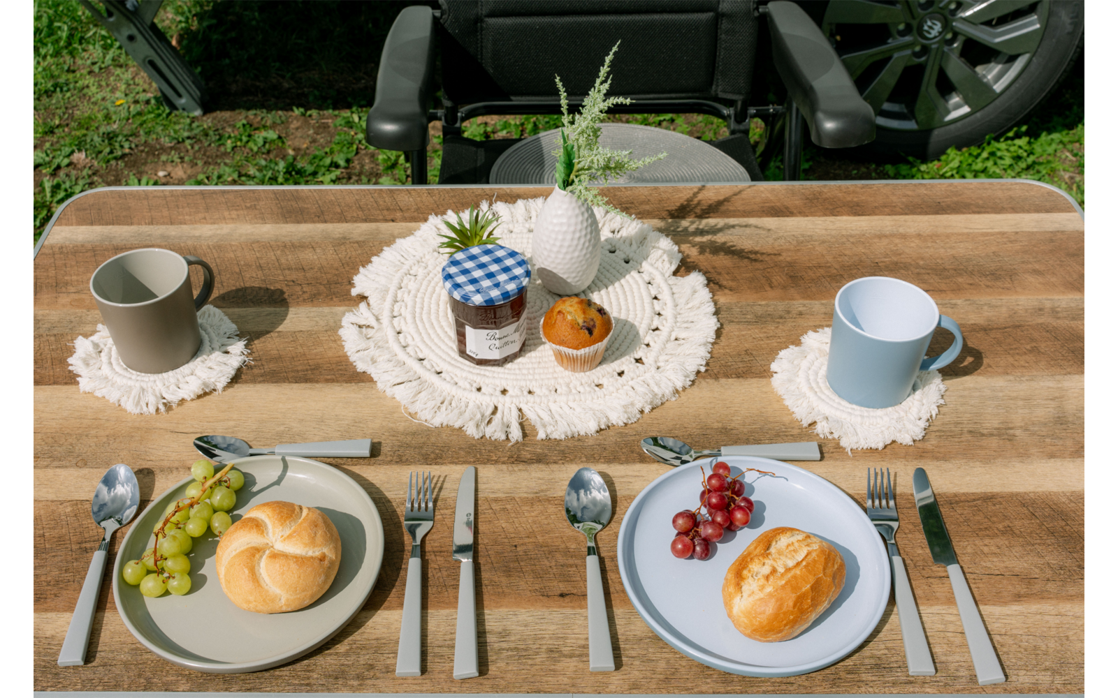 Houten tafel met twee gedekte plaatsen: links een licht bord met een broodje en groene druiven, rechts een blauw bord met een broodje en rode druiven; vorken links, messen en lepels rechts; twee mokken op gehaakte onderzetters; in het midden een gehaakt kleedje met een jampot, een witte keramische ananas en een oranje vrucht; gras en autowiel op de achtergrond