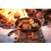 Gusseiserne Pfanne mit Steak und halbierten Zwiebeln auf Holzscheiten, Flammen und Schnee im Hintergrund