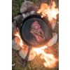 Gusseiserne Grillpfanne über offenem Feuer mit einem rohen Steak und einem Zweig Rosmarin; Pfannenstiel, Steine und Gras sichtbar