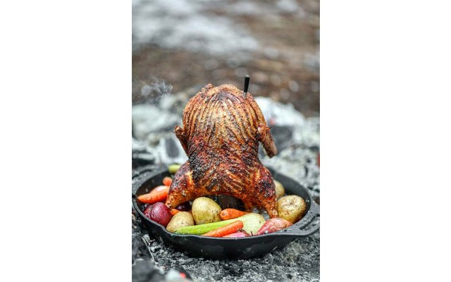 Ganzes Brathähnchen mit knuspriger Haut, stehend in Gusseisenpfanne mit Kartoffeln und Karotten, Metallspieß oben, Rauch im Hintergrund