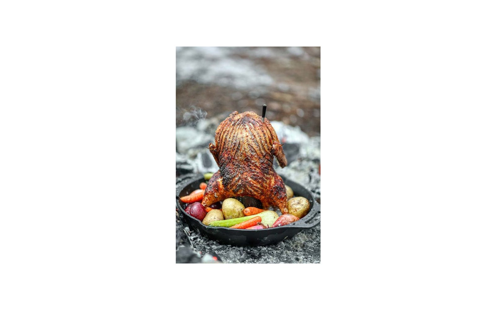 Ganzes Brathähnchen mit knuspriger Haut, stehend in Gusseisenpfanne mit Kartoffeln und Karotten, Metallspieß oben, Rauch im Hintergrund