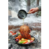 Ganzes Brathähnchen aufrecht in einer gusseisernen Pfanne mit Kartoffeln, Karotten und Zwiebeln; eine Hand gießt aus einem kleinen Topf Bratensaft darüber; Glut und verschneiter Wald im Hintergrund