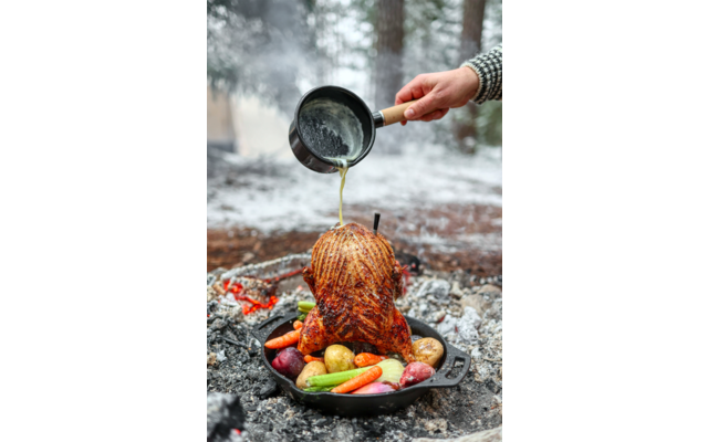 Ganzes Brathähnchen aufrecht in einer gusseisernen Pfanne mit Kartoffeln, Karotten und Zwiebeln; eine Hand gießt aus einem kleinen Topf Bratensaft darüber; Glut und verschneiter Wald im Hintergrund