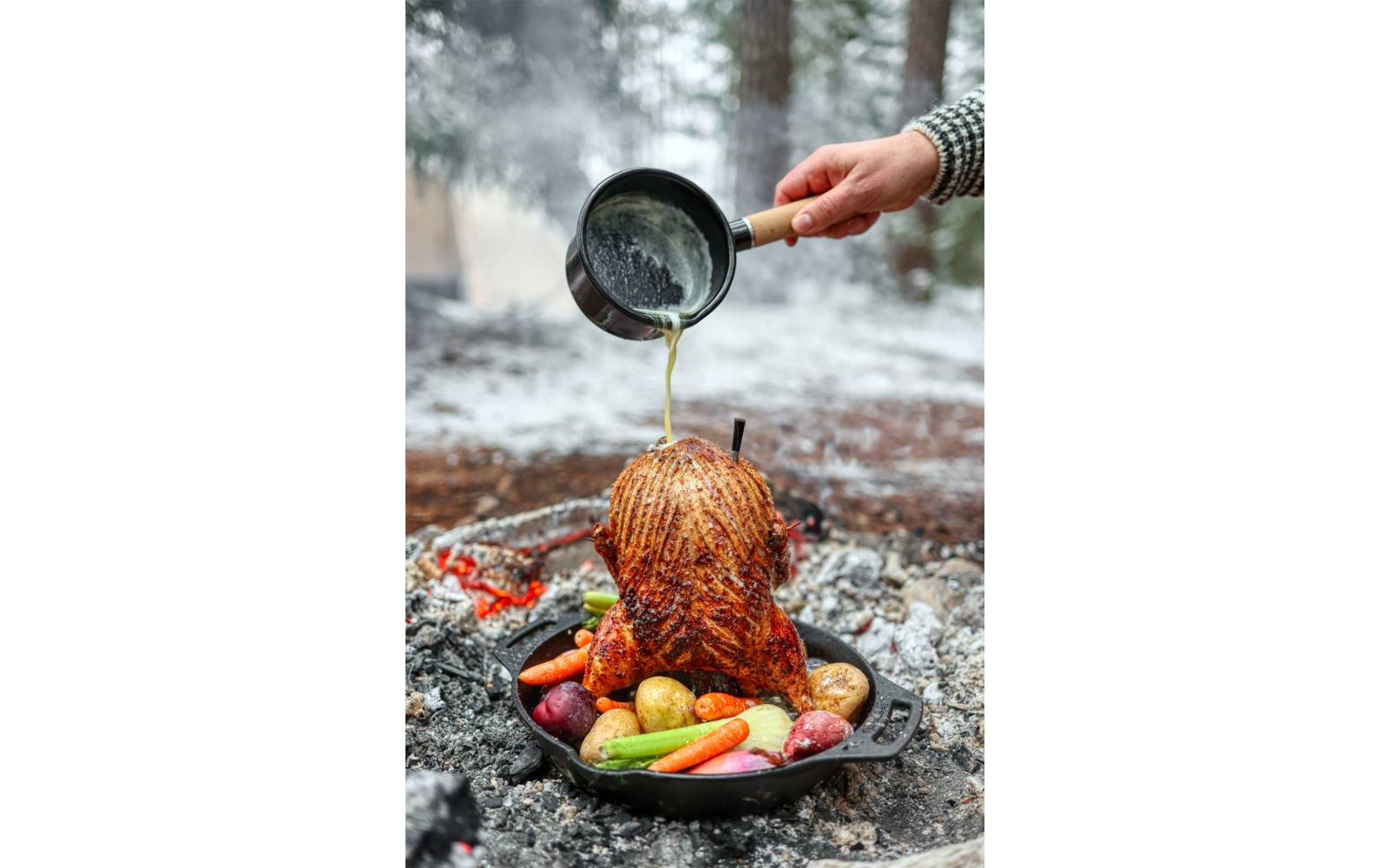 Ganzes Brathähnchen aufrecht in einer gusseisernen Pfanne mit Kartoffeln, Karotten und Zwiebeln; eine Hand gießt aus einem kleinen Topf Bratensaft darüber; Glut und verschneiter Wald im Hintergrund