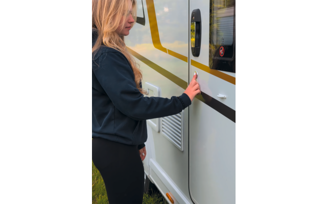 Person mit langen blonden Haaren in dunkler Jacke und dunkler Hose berührt den Türgriff eines weißen Wohnmobils mit gelb-schwarzem Streifen; Lüftungsgitter, kleines Fenster mit Aufkleber und Gras sind sichtbar
