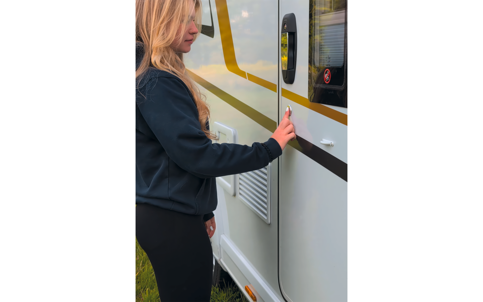Person mit langen blonden Haaren in dunkler Jacke und dunkler Hose berührt den Türgriff eines weißen Wohnmobils mit gelb-schwarzem Streifen; Lüftungsgitter, kleines Fenster mit Aufkleber und Gras sind sichtbar