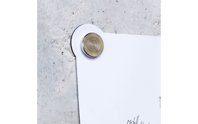 Weiße Ecke eines Blatts mit abgerundeter Kante, oben links mit rundem goldfarbenem Magnet an grauer Betonwand befestigt; unten rechts teilweise Handschrift sichtbar
