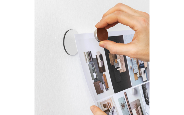 Hand hält einen runden Magneten, befestigt ein Blatt mit Bildern von Holzmustern an einer weißen Wand