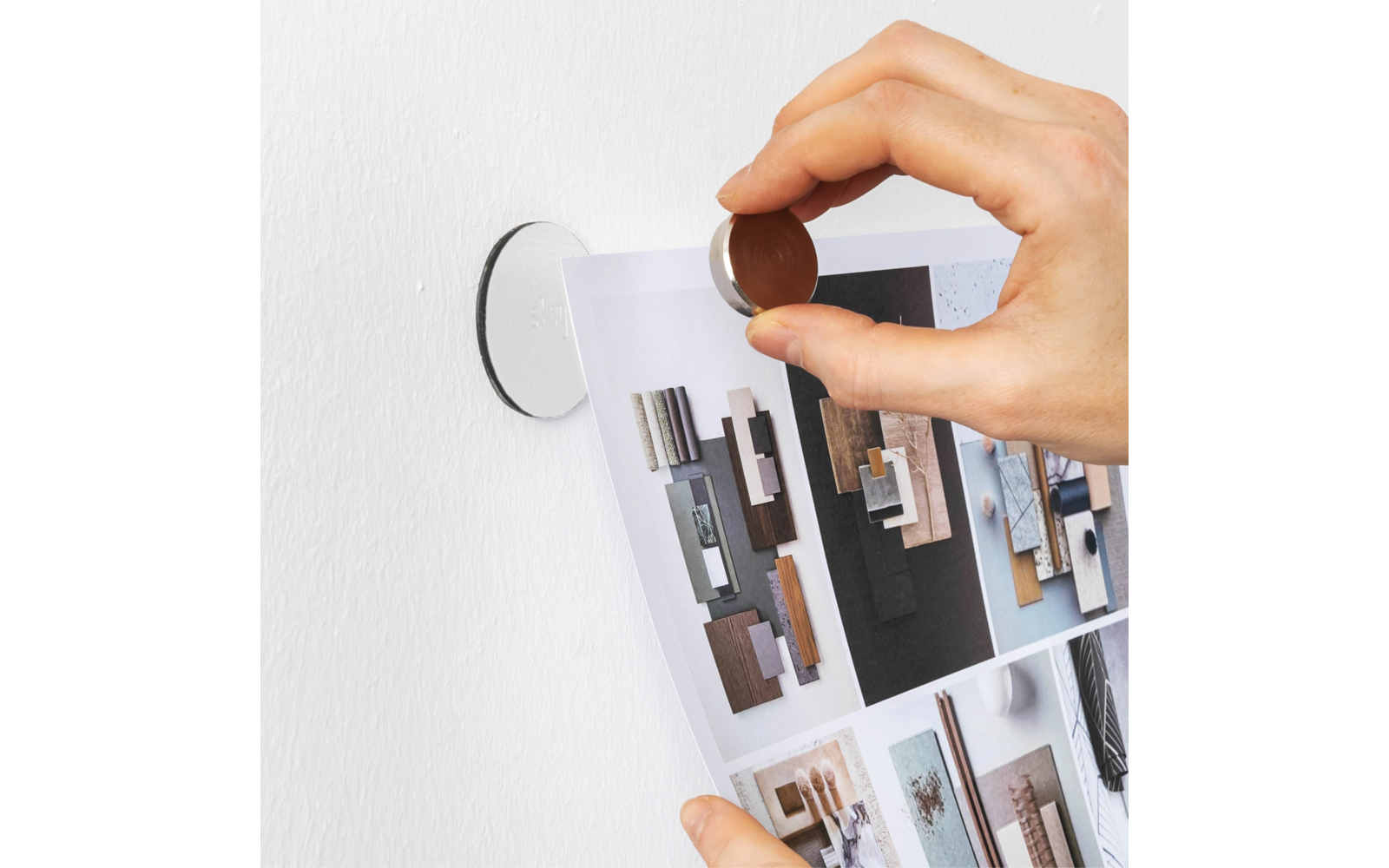 Hand hält einen runden Magneten, befestigt ein Blatt mit Bildern von Holzmustern an einer weißen Wand