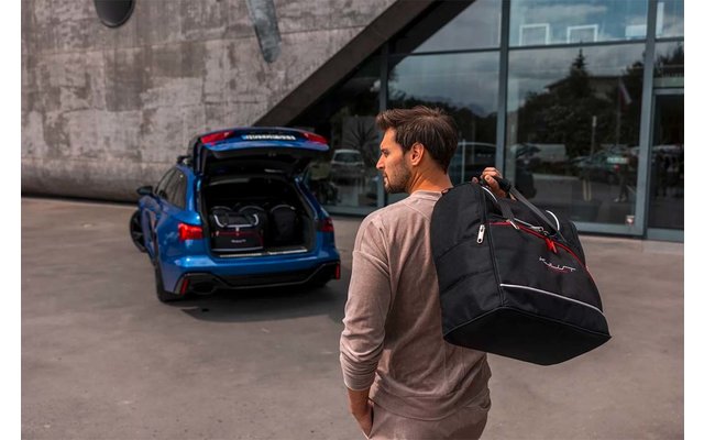Mann von hinten trägt schwarze Reisetasche über der Schulter und geht auf den geöffneten Kofferraum eines blauen Autos vor einem Gebäude mit Glasfassade zu