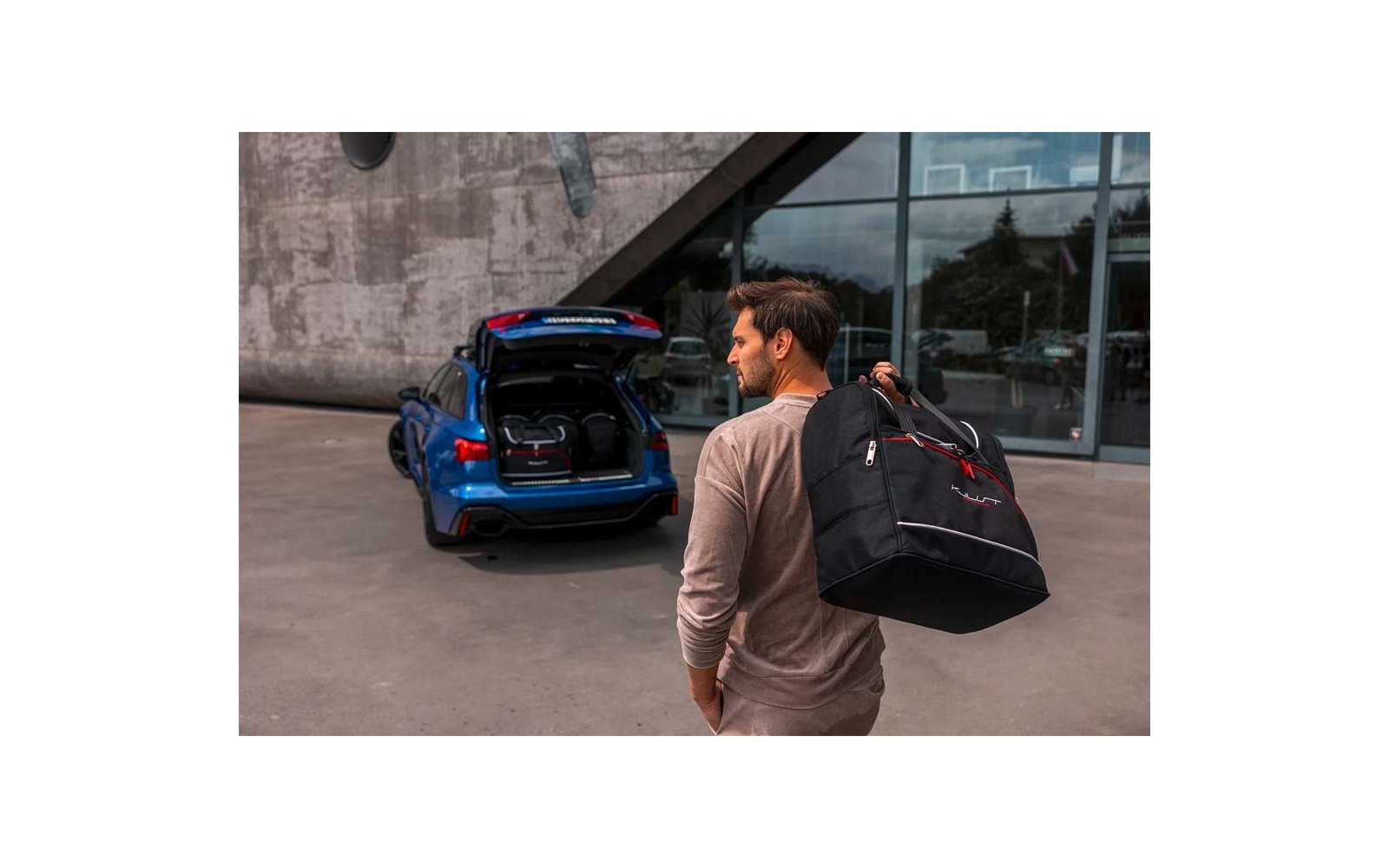 Mann von hinten trägt schwarze Reisetasche über der Schulter und geht auf den geöffneten Kofferraum eines blauen Autos vor einem Gebäude mit Glasfassade zu