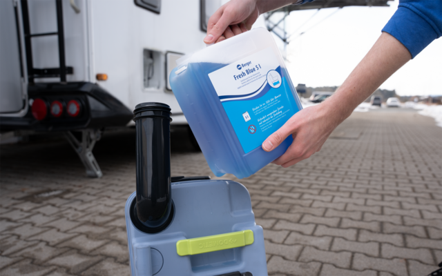 Hände halten einen blauen Kunststoffkanister und gießen eine blaue Flüssigkeit in einen grauen Kunststoffbehälter mit schwarzem Einfüllstutzen; links ein Wohnwagen, gepflasterter Boden