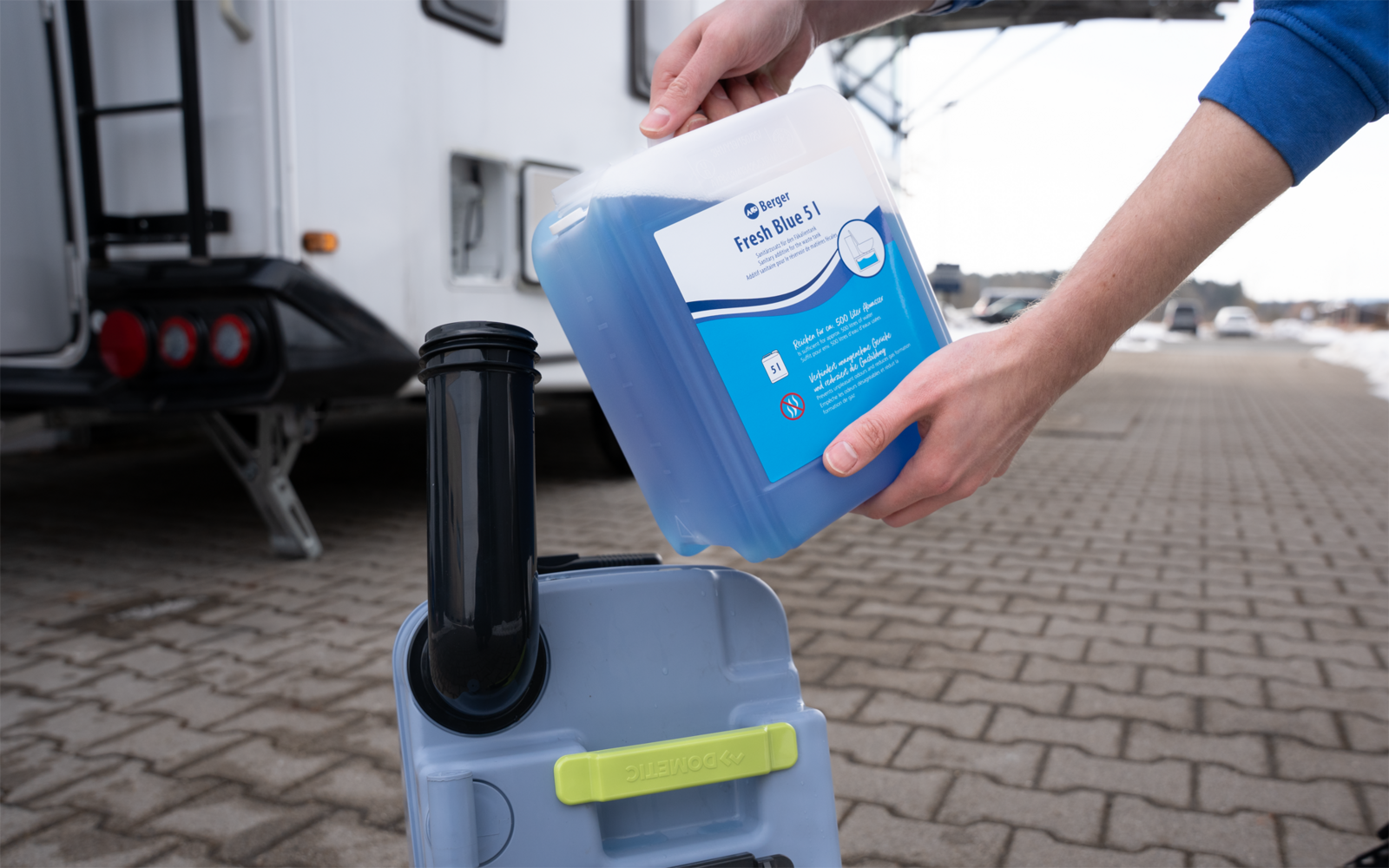 Hände halten einen blauen Kunststoffkanister und gießen eine blaue Flüssigkeit in einen grauen Kunststoffbehälter mit schwarzem Einfüllstutzen; links ein Wohnwagen, gepflasterter Boden