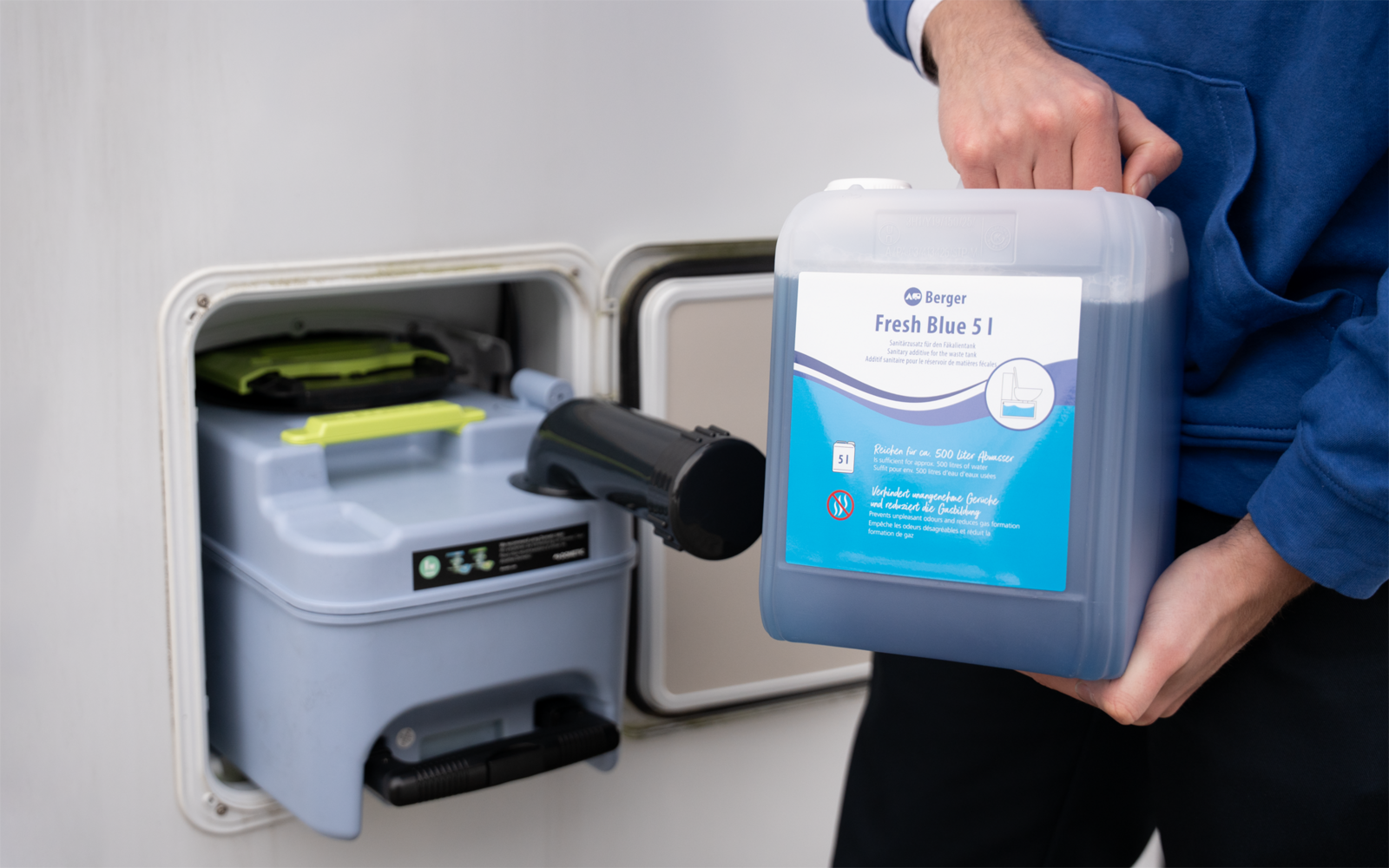Person in blauem Oberteil hält einen blauen 5‑Liter-Kanister mit Etikett 'Fresh Blue 5 L' neben einem offenen Klappfach in einer weißen Außenwand, darin eine graue Kassette mit grünem Griff