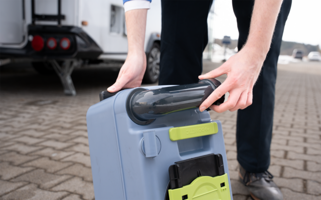 Hände einer Person drehen einen schwarzen Schraubverschluss an einem grauen Kunststoffbehälter mit grünem Verschluss auf gepflastertem Boden neben einem Wohnwagen