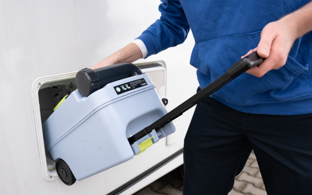 Person in blauem Oberteil zieht ein graues, rollbares Gerät mit schwarzem Griff und digitalem Display aus dem offenen Seitenfach eines weißen Wohnwagens; Pflasterboden sichtbar