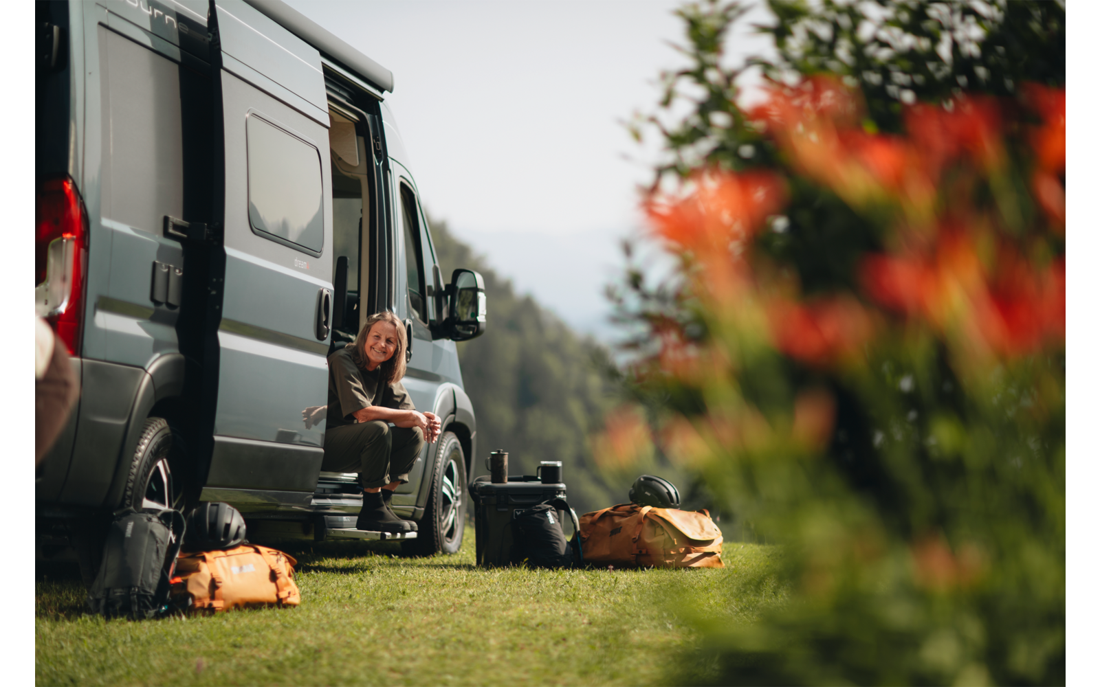 Persona sentada en la puerta lateral abierta de una furgoneta camper sobre césped; mochilas y equipo en el suelo delante; flores rojas desenfocadas en primer plano; montañas y árboles al fondo