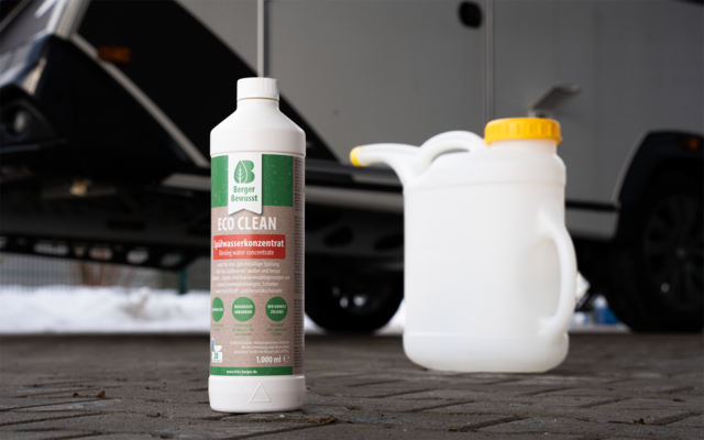 Weiße Kunststoffflasche mit grünem Etikett "ECO CLEAN" auf gepflastertem Hof, daneben ein weißer Kunststoffkanister mit gelbem Deckel; im Hintergrund zwei geparkte Autos und Schneereste