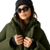 Person mit braunen Haaren, schwarze Mütze und Sonnenbrille, olivgrüne Steppjacke, hält die Jacke am Reißverschluss