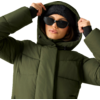 Person mit grüner gesteppter Winterjacke mit Kapuze, schwarzer Mütze und schwarzer Sonnenbrille, hält die Kapuze mit beiden Händen