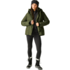 Person steht, trägt grüne gesteppte Winterjacke mit Kapuze, schwarze Strickmütze, schwarzen Reißverschluss-Pullover, schwarze Leggings und schwarze Winterstiefel mit weißem Fellbesatz; Hände halten die Jackenkanten