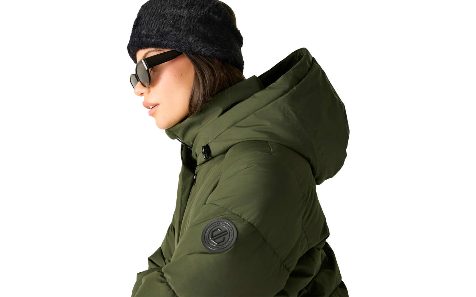 Person im Profil mit dunkelgrauer Strickmütze, schwarzer Sonnenbrille und olivgrüner Steppjacke mit Kapuze und rundem Ärmelpatch