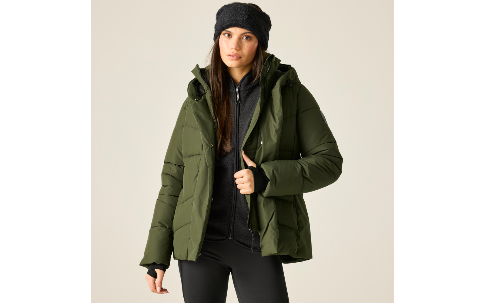 Frau in olivgrüner gesteppter Jacke, schwarzer Mütze, schwarzem Reißverschluss-Oberteil und schwarzen Leggings vor hellem Hintergrund