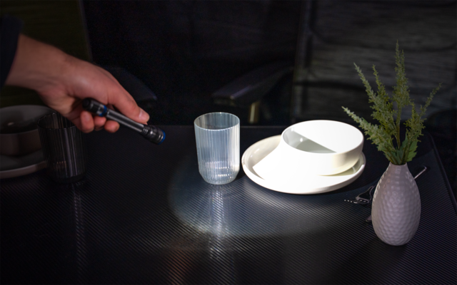 Hand mit Taschenlampe leuchtet auf einen transparenten Becher, gestapelte weiße Schalen auf einem Teller und ein kleines Väschen mit grünen Zweigen auf einem dunklen Tisch