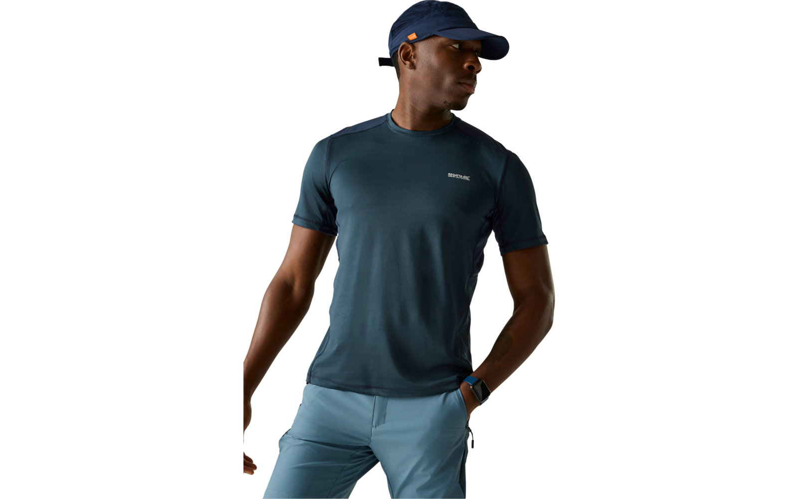 Person mit dunkelblauer Cap, dunkeltürkisem T‑Shirt und hellblauen Hosen, steht vor neutralem Hintergrund, schaut zur Seite, eine Hand in der Hosentasche