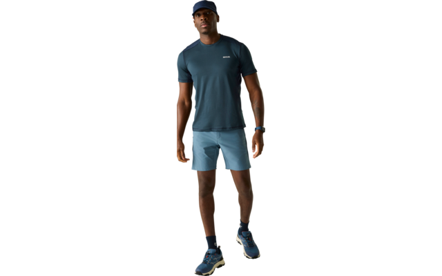 Person steht auf hellem Hintergrund, trägt dunkle Kappe, dunkelblaues T‑Shirt mit kleinem Brustlogo, hellblaue Shorts, dunkle Socken und blaue Laufschuhe